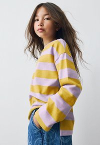 Jeune personne avec de longs cheveux portant un pull rayé jaune et lavande et un jean bleu sur un fond blanc uni.