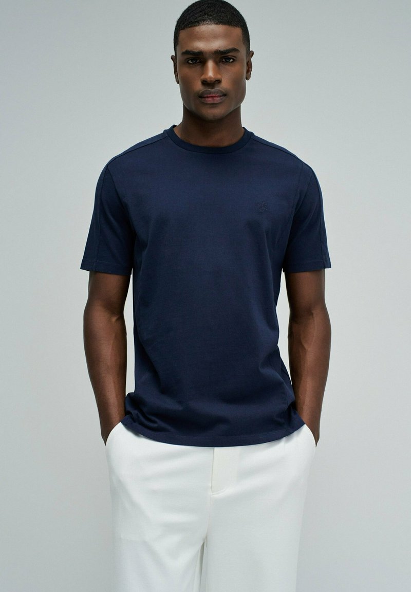 Camiseta azul marino de algodón con mangas cortas, cuello redondo y detalle sutil de logo en el pecho, combinada con pantalones blancos.