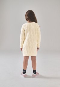 Megztinė su kremine suknelė su trijų kvadratų tekstūriniu raštu, su ribotu rankogaliu ir apačia. Derinama su rožiniais sportbačiais ir juodomis kojinėmis.