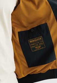 Poche à boutons noirs étiquetée "Winglock" sur un tissu en mesh jaune moutarde de la doublure intérieure d'une veste, avec un tissu extérieur blanc visible.