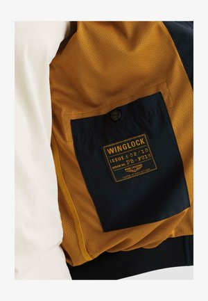 Poche à boutons noirs étiquetée "Winglock" sur un tissu en mesh jaune moutarde de la doublure intérieure d'une veste, avec un tissu extérieur blanc visible.