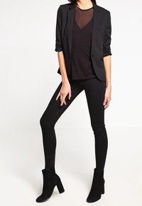 Blazer nero aderente con maniche arrotolate, indossato sopra una maglietta nera trasparente, abbinato a jeans skinny neri e stivaletti neri.