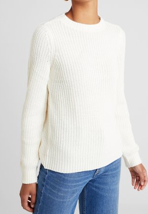 Vrouw die een wit gebreide trui met lange mouwen draagt, gecombineerd met een blauwe spijkerbroek, tegen een effen achtergrond.