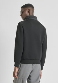 Sudadera negra con cuello alto, tejido texturizado y puños acanalados. La parte trasera presenta un delicado detalle de costura. Pantalones grises visibles en la parte inferior.