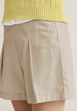 Beige knælange shorts med frontplisseret, båret med en gul ribstrikket sweater og en hvid skjorte under.
