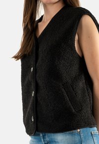 Chaleco negro sin mangas de forro polar texturizado con diseño de cuello en V, que cuenta con cierres de botón plateados y un bolsillo lateral.