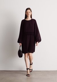 Čierne plisované šaty s dlhými rukávmi a smockovaným páse. Modelka drží malú čiernu kabelku a má na sebe šľapky na opätku.