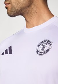 Baltas sportinis marškinėlis su juodomis Adidas juostomis ir Mančesterio Unitedo logotipu. Medžiaga atrodo lygi ir lengva, su apvalia iškirpte.