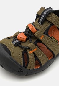Olivengrønn sandal med svarte detaljer, med gummisåle, justerbare snører og "VASKBAR"-merke. Teksturen er myk stoff med utskjæringer.