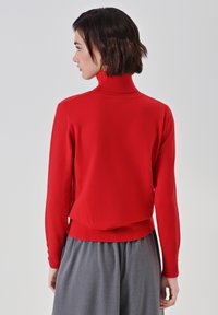 Roter Rollkragenpullover mit taillierter Passform, gerippter Textur und langen Ärmeln. Besitzt eine verkürzte Länge und Knopfdetails an den Ärmeln.