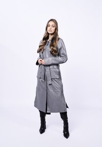 Trench coat in pelle grigia con vita cintrata, chiusura a bottoni, due tasche frontali e design lungo fino al ginocchio. Texture lucida e vestibilità sartoriale.