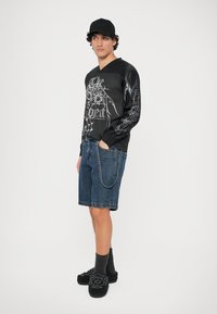 Black long-sleeve shirt with graphic print, denim shorts, black slippers, and gray socks. Accessories include a black cap and chain detail.