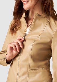 Beige leren jack met een hoge kraag, gladde textuur en een ritssluiting aan de voorkant. Heeft zijzakken en stikseldetails op het lichaam.