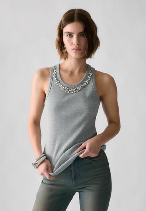 Vrouw met kort bruin haar in een grijs hemdje met kristallen versieringen en blauwe jeans staat naar voren gericht en houdt de zoom van haar shirt vast.