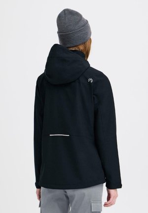 Person som bærer svart hettejakke med lite logo på skulderen og grå lue, sett bakfra mot hvit bakgrunn.