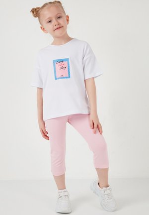 Jeune fille aux cheveux blonds coiffés en chignons, portant un T-shirt blanc avec le texte « Every love story », un legging capri rose et des baskets blanches, debout sur un sol blanc.