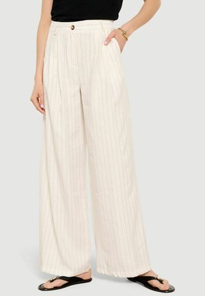 Kobieta w kremowych, szerokich spodniach z wysokim stanem i cienkimi pionowymi paskami, czarna bluzka, złota bransoletka i czarne sandały.