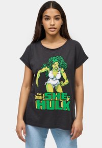 Camiseta de manga corta gris oscuro hecha de algodón, con un gráfico verde y amarillo de She-Hulk y un texto en negrita debajo.