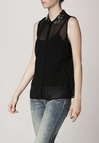 Top nero senza maniche in tessuto trasparente, con un colletto metallico testurizzato. Abbinato a jeans chiari strappati.