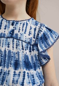 Blå och vit mönstrad topp med knäppning fram, volangärmar med scallopdetaljer och textil med en akvarelleffekt.