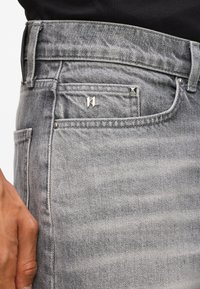 Sivé deninové džínsy s stredným pásom, vreckom so strieborným logom a dekoratívnymi striebornými cvokmi v blízkosti pásu.