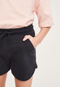Schwarze Baumwollshorts mit geripptem Bund, Kordelzug, abgerundetem Saum und Seitentaschen, kombiniert mit einem hellrosa Shirt.