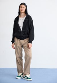 Sudadera negra con cremallera, capucha, bolsillos frontales y detalles de costura ligera, combinada con pantalones beige y zapatillas verdes.