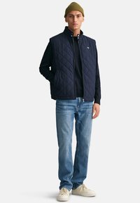 Navy quilted vest, zip closure, high collar, worn over a black sweater. Paired with light blue jeans and cream sneakers. Olive beanie on head.
