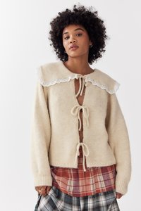 Vrouw met krullend haar die een beige gebreide cardigan met koorden en kantenkraag draagt over een rood en blauw geruit rokje tegen een witte achtergrond.