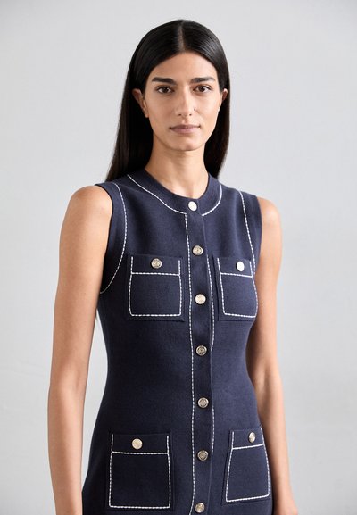 Mouwloze marineblauwe jurk met witte stiksel details, voorzien van twee voorzakken en metalen knopen aan de voorkant. Gladde textuur.
