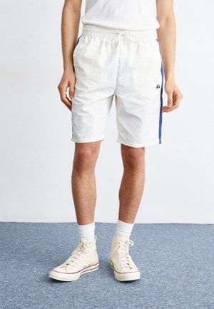 Person trägt weiße sports Shorts mit blauen Seitenstreifen, weiße Socken und cremefarbene High-Top-Sneaker und steht auf einem blauen Teppich.