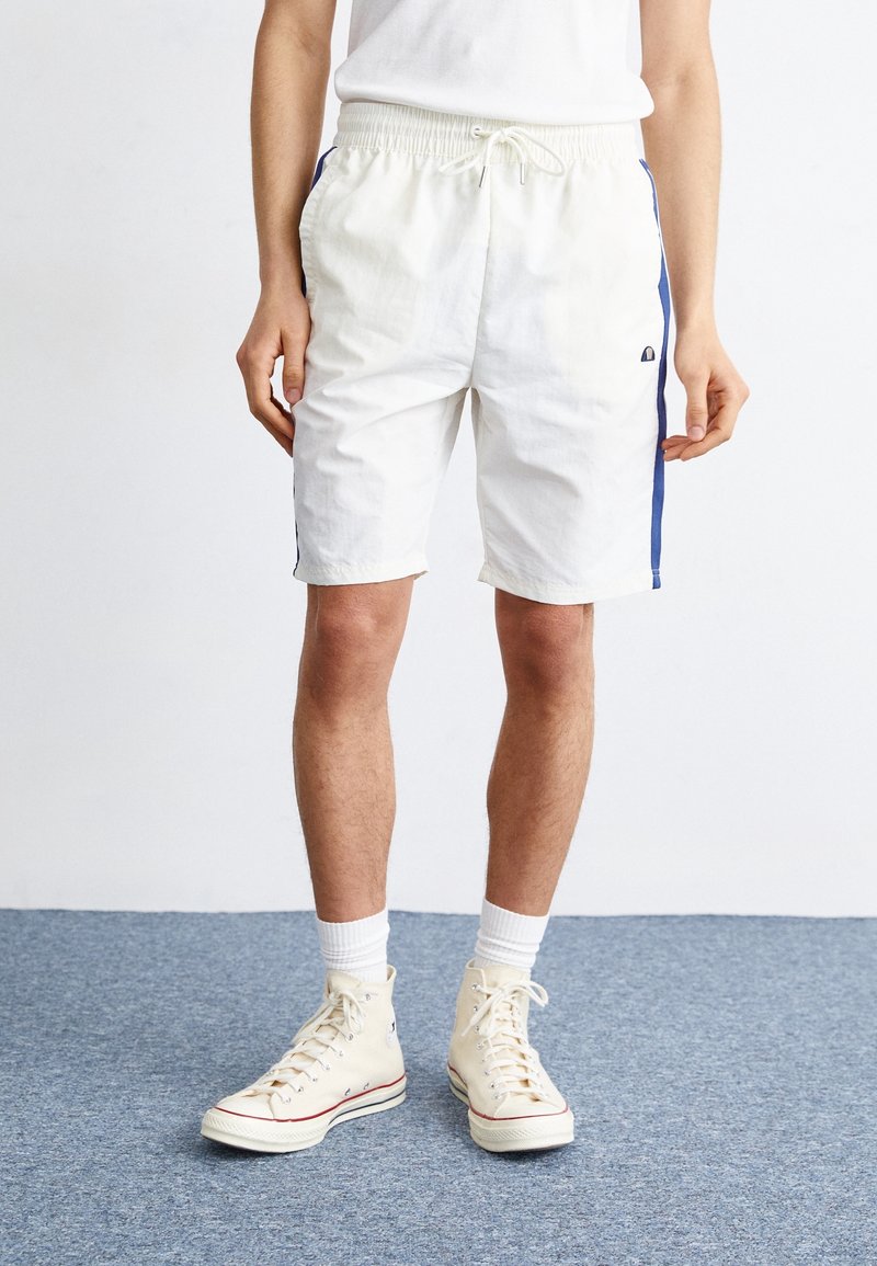 Personne portant un short de sport blanc avec des bandes latérales bleues, des chaussettes blanches et des baskets montantes écrues, debout sur un tapis bleu.