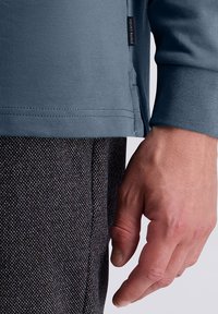 Primo piano della mano di una persona accanto all'anca, che indossa un top blu a maniche lunghe con un'etichetta "pierre cardin" e pantaloni scuri con texture.