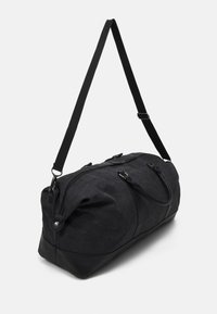 Sac de sport noir en tissu texturé avec des accents en cuir. Dispose de deux poignées supérieures et d'une bandoulière amovible pour un transport polyvalent.