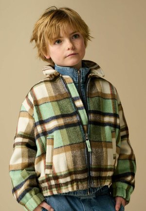 Enfant aux cheveux courts châtain clair portant une veste zippée à carreaux verts, marron et blancs sur une veste en jean, devant un fond beige.