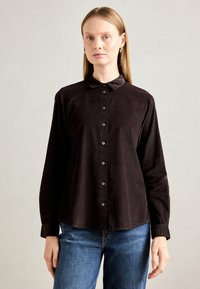 Woman with straight blonde hair wearing gold hoop earrings, a black buttoned long-sleeve shirt, and blue jeans against a plain background.