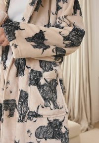 Személy bézs polár köntöst visel fekete hegyvidéki tehenek skicceivel, az ujját igazítja, beltéren, krémszínű függöny háttér előtt.