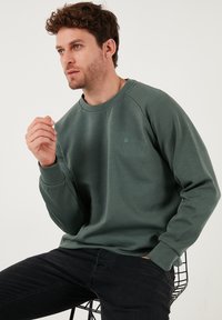 Zelený pulóver s okrúhlym výstrihom, dlhými rukávmi a malým logom na hrudi. Čierne džínsy a drôtová stolička v pozadí.