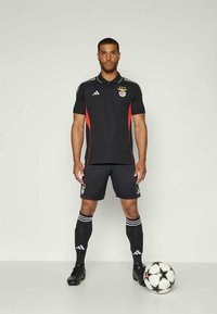 Svart Adidas fotballuniform med kortermede ermer, røde detaljer og en klubbemblem. Idrettsutøveren står ved siden av en svart og hvit fotball.