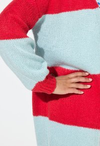 Strickjacke mit roten und hellblauen Streifen, langärmlig und aus strukturiertem Stoff. Enthält einen auffälligen roten Ärmelbund.