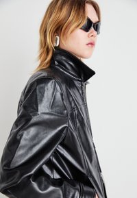 Sort faux læderjakke med høj krave, med lynlås og blank overflade. Båret med mørke solbriller og store sølvsmykker.