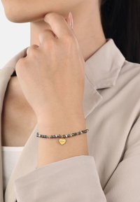 Bracciale di perline con perline nere e dorate, con un ciondolo a forma di cuore dorato inciso con la parola "amor". Indossato su un polso.