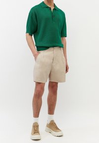 Polo verde a maniche corte con colletto e dettagli con bottoni, abbinato a pantaloni corti beige e sneakers di colore chiaro. Tessuto di superficie liscia.