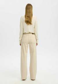 Femme aux cheveux longs de dos, portant un pull crème et un pantalon beige à jambes larges, sur un fond blanc uni.