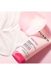 Crème pour les mains Nuxe Very Rose dans un tube rose doux avec un capuchon rose à bascule. Le texte indique "Crème pour les mains apaisante avec extrait de rose." La texture crémeuse est visible.