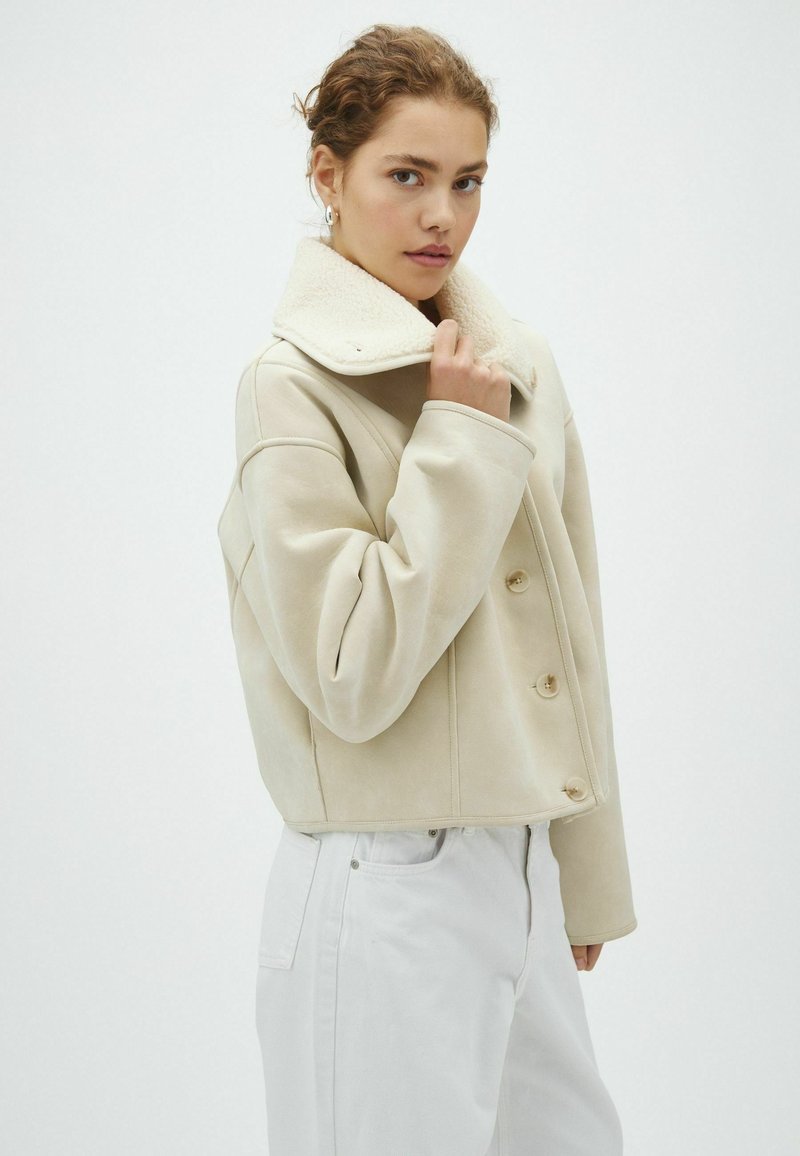 Jeune femme aux cheveux bouclés portant une veste beige en shearling et un pantalon blanc, tenant le col de la veste d'une main.