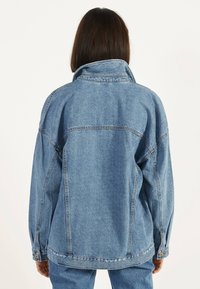 Blå denimjacka med hög krage och oversized passform. Har synliga sömmar och knappstängning vid ärmsluten. Klassisk textur.