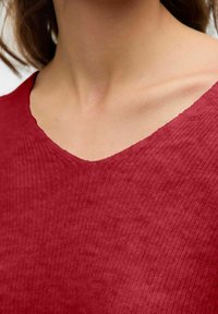 Haut côtelé rouge avec un décolleté en V. Le tissu a une texture douce, mettant en valeur les motifs subtils créés par les côtes.