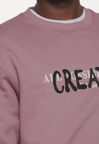 Sweatshirt rose avec un col rond et des lettres texturées. Présente "CREATIVE" en noir et un texte blanc plus petit au-dessus. Tissu doux.