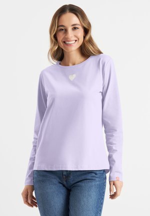 Uśmiechnięta kobieta z falującymi włosami ubrana w lawendową bluzkę z długim rękawem z małym białym sercem na piersi oraz niebieskie dżinsy na białym tle.